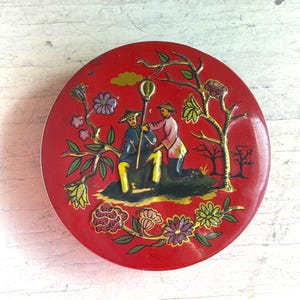 May include: A round red metal tin featuring a colorful illustration of two figures wearing conical hats, one seated and one standing, holding a tall staff. The design includes stylized trees, blooming flowers, and a small yellow cloud on a red background.