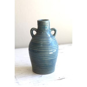 May include: A blue ceramic vase with two small handles and a textured, horizontal striped pattern. The vase has a rounded body and a narrow neck. The color is a muted blue-green, suitable for home decor.
