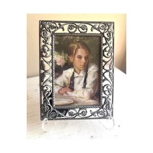 May include: A rectangular picture frame with a dark grey metallic finish and an intricate, raised leaf and vine pattern. The frame holds an oil painting of a person with light hair wearing a white shirt with dark straps, seated at a table.