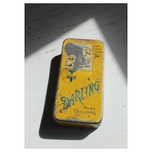 May include: A rectangular, vintage tin with a yellow background and the word "DARLING" in teal script. The tin has a distressed appearance with a faded image and text, including "Hors Concours."