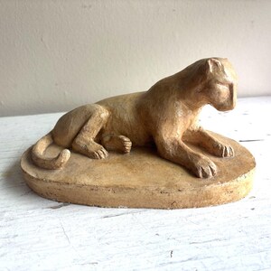 May include: A tan-colored sculpture of a large cat in a reclining position. The feline figure is depicted lying down with its front paws extended forward and its tail curled near its hind legs, all resting on an oval-shaped base of the same color.