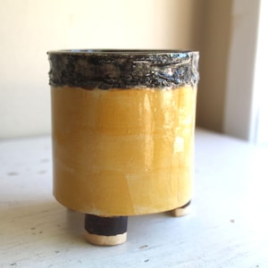 May include: A cylindrical ceramic planter with a yellow body and a textured, dark brown rim. The planter sits on three short legs with a dark brown base and a cream-colored bottom. The planter is suitable for small plants.