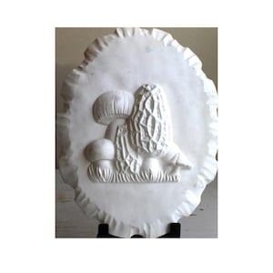 May include: White ceramic wall plaque with a raised relief design of mushrooms. The plaque has a scalloped edge and is mounted on a black stand.