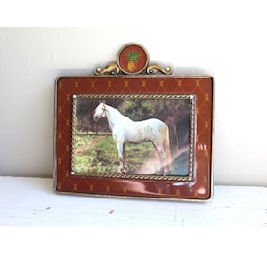 Puede incluir: Un marco de fotos rectangular con una pintura de un caballo blanco en un campo. El marco es marrón con un borde dorado y un emblema de piña en la parte superior. El marco mide aproximadamente 20 cm de ancho y 15 cm de alto.