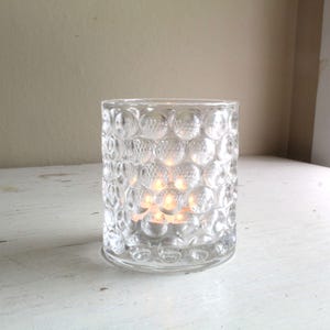 May include: A clear glass candle holder with a textured, bubble-like design. The holder is lit with a tea light candle.