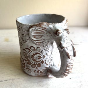 Könnte beinhalten: Ein Keramikbecher mit weißer Glasur und braunen floralen Reliefmustern. Der Henkel ist als Elefantenkopf mit großen Ohren, Augen und einem Rüssel geformt, der sich nach unten biegt und den Griff des Bechers bildet.