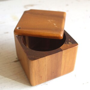 May include: A square wooden box with a circular interior cutout and a matching square lid. The box is crafted from medium-toned brown wood with visible grain patterns. The lid is partially open, revealing the hollow, cylindrical centre of the container.