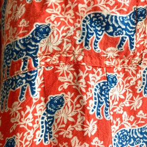May include: Close-up of a fabric with a repeating pattern of blue tigers and white floral elements on a red background. The fabric appears to be cotton or a similar material, with visible seams.