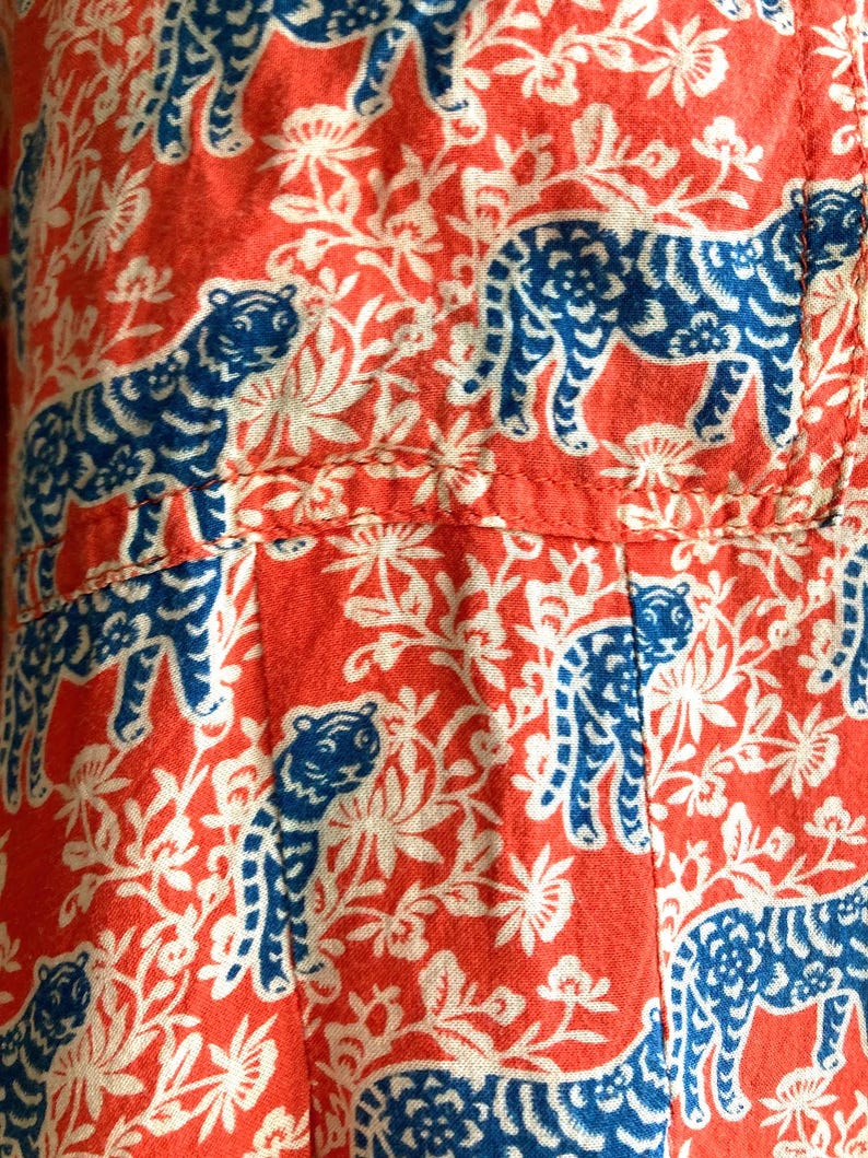 May include: Close-up of a fabric with a repeating pattern of blue tigers and white floral elements on a red background. The fabric appears to be cotton or a similar material, with visible seams.