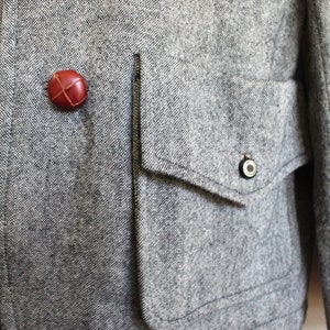 May include: A grey tweed jacket with a large flap pocket and two brown leather buttons. The pocket has a small metal button on the flap.