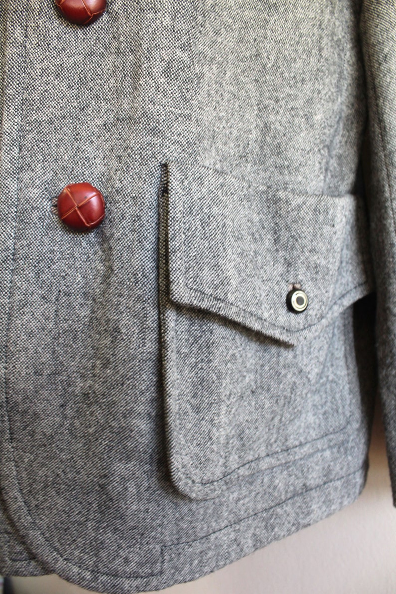 May include: A grey tweed jacket with a large flap pocket and two brown leather buttons. The pocket has a small metal button on the flap.