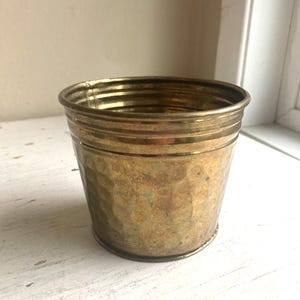 May include: A small, hammered brass planter with a cylindrical shape. The planter has a textured surface and a slightly flared rim. The brass has a warm, aged patina, giving it a vintage look. Ideal for small plants or as a decorative accent.