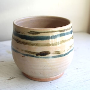 Puede incluir: Un vaso de cerámica hecho a mano con base beige y rayas horizontales en verde azulado, marrón y beige. La taza tiene forma redondeada y base color terracota. El interior es de color beige claro.