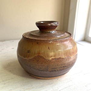 May include: A round ceramic sugar bowl with a lid, featuring a glaze that transitions from a dark brown base to a lighter tan and golden yellow upper section. The lid has a small, raised central knob handle and a smooth, glossy finish.