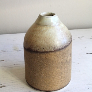 Puede incluir: Un pequeño jarrón de cerámica con un esmalte marrón claro en la mitad inferior y un esmalte blanco en la mitad superior. El jarrón tiene un cuerpo redondeado y una abertura estrecha.