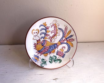 May include: A small, white ceramic plate with a hand-painted design of a colorful bird with a long tail. The bird is surrounded by floral motifs in shades of pink, yellow, and blue.