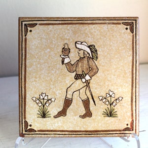 Pode incluir: Um azulejo de cerâmica vintage com uma borda marrom e um fundo bege. O azulejo apresenta uma ilustração pintada à mão de uma pessoa com um chapéu segurando um pássaro. A pessoa está vestindo uma túnica e calças marrons. O azulejo também apresenta um design floral com flores brancas.