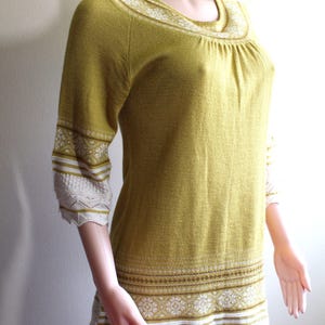 Pode incluir: Um suéter de malha amarelo com detalhes em branco e marrom. O suéter tem um decote redondo e mangas 3/4. As mangas e a bainha inferior têm uma renda branca.