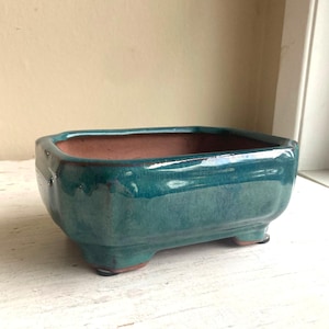 May include: A rectangular bonsai pot with a glossy teal glaze. The ceramic pot has a brown interior and sits on three small feet. The pot's shape is slightly curved, and the edges are rounded.