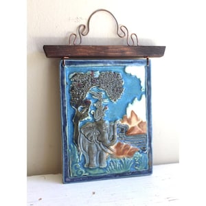 May include: A rectangular ceramic wall hanging with a blue background and a relief image of an elephant, tree, and figures. The piece has a dark wood top and copper wire hanger. The artwork is framed with a dark blue border.