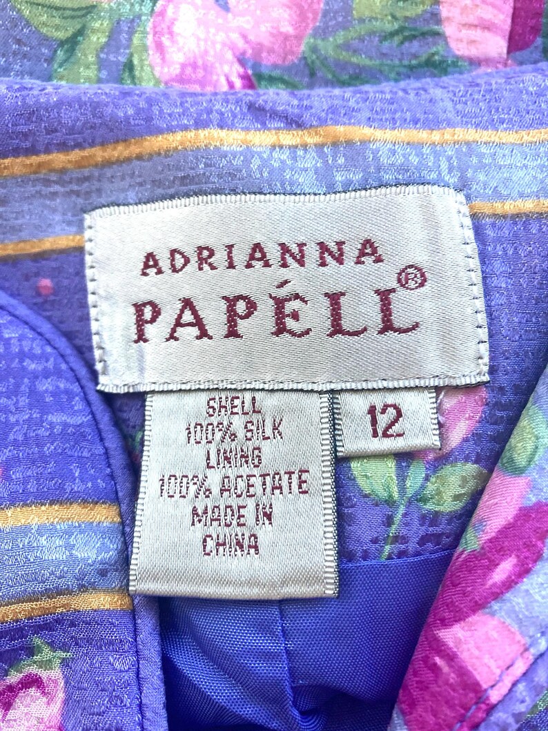 May include: Close-up of a label from an Adrianna Papell garment. The label is white with the brand name in burgundy. The label indicates the shell is 100% silk, the lining is 100% acetate, and it is made in China. The size is 12.