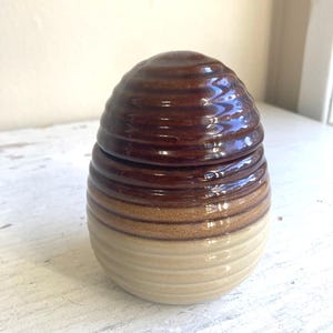May include: A ceramic jar with a lid. The jar is a light beige color at the base, transitioning to a medium brown in the middle, and a dark brown at the top. The jar has a ribbed texture.