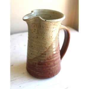 May include: A handmade ceramic pitcher with a tan and reddish-brown color scheme. The pitcher has a curved spout and a handle. The upper portion is tan with dark speckles, transitioning to reddish-brown at the base. The pitcher is likely used for serving beverages.