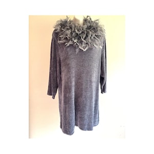 May include: A long-sleeved grey tunic featuring a textured fabric and a decorative collar made of grey feathers. The garment has a straight hemline and a loose fit, with the feather trim encircling the neckline in a dense, layered arrangement.