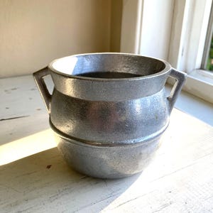May include: A small, silver-colored metal cauldron with two handles. The cauldron has a rounded base and a wider top, with a textured surface. It sits on a white, weathered surface near a window.