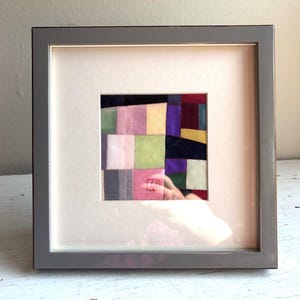 Puede incluir: Una pieza de arte textil enmarcada de forma cuadrada que presenta un mosaico de pequeños cuadrados de tela multicolores en tonos rosa, amarillo, verde, morado y rojo oscuro. La obra está bajo cristal en un marco gris liso con un paspartú crema.