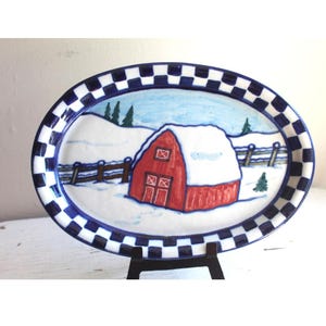 May include: Oval ceramic serving platter with a winter scene. The platter features a red barn with a snow-covered roof, set against a snowy landscape and blue sky. The rim has a blue and white checkered pattern.