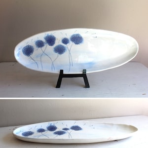 Op de afbeelding: Witte ovale serveerschaal van keramiek met een blauw en wit paardenbloemmotief. De schaal staat op een zwarte standaard.