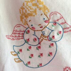 May include: Embroidered white fabric with a cross-stitch design of an angel with yellow hair, pink wings, and a blue dress with pink and green flowers. The angel is holding a small object in its hands.