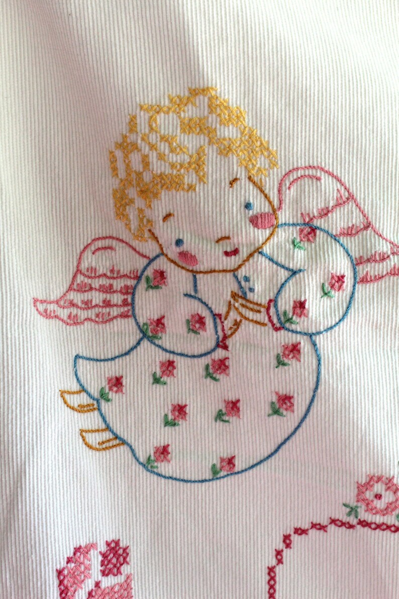 May include: Embroidered white fabric with a cross-stitch design of an angel with yellow hair, pink wings, and a blue dress with pink and green flowers. The angel is holding a small object in its hands.