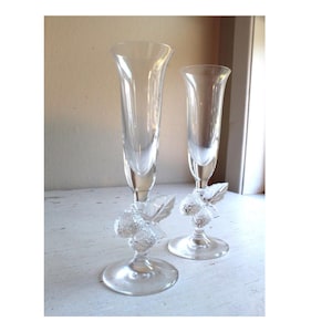 May include: Two clear glass champagne flutes with a detailed berry design on the base. The flutes are tall and slender with a delicate stem.