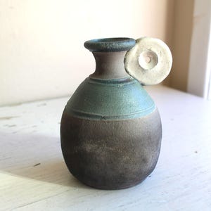 Könnte beinhalten: Eine kleine, runde Keramikvase mit brauner und grüner Glasur. Die Vase hat einen einzigartigen Griff, der aus einer weißen, runden Scheibe mit einem Loch in der Mitte besteht.