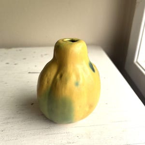 May include: A small, bulbous ceramic vase with a smooth, matte finish. The surface features a mottled color palette of warm yellow, soft green, and subtle dark blue accents. The vase has a narrow, circular opening at the top and a rounded body.