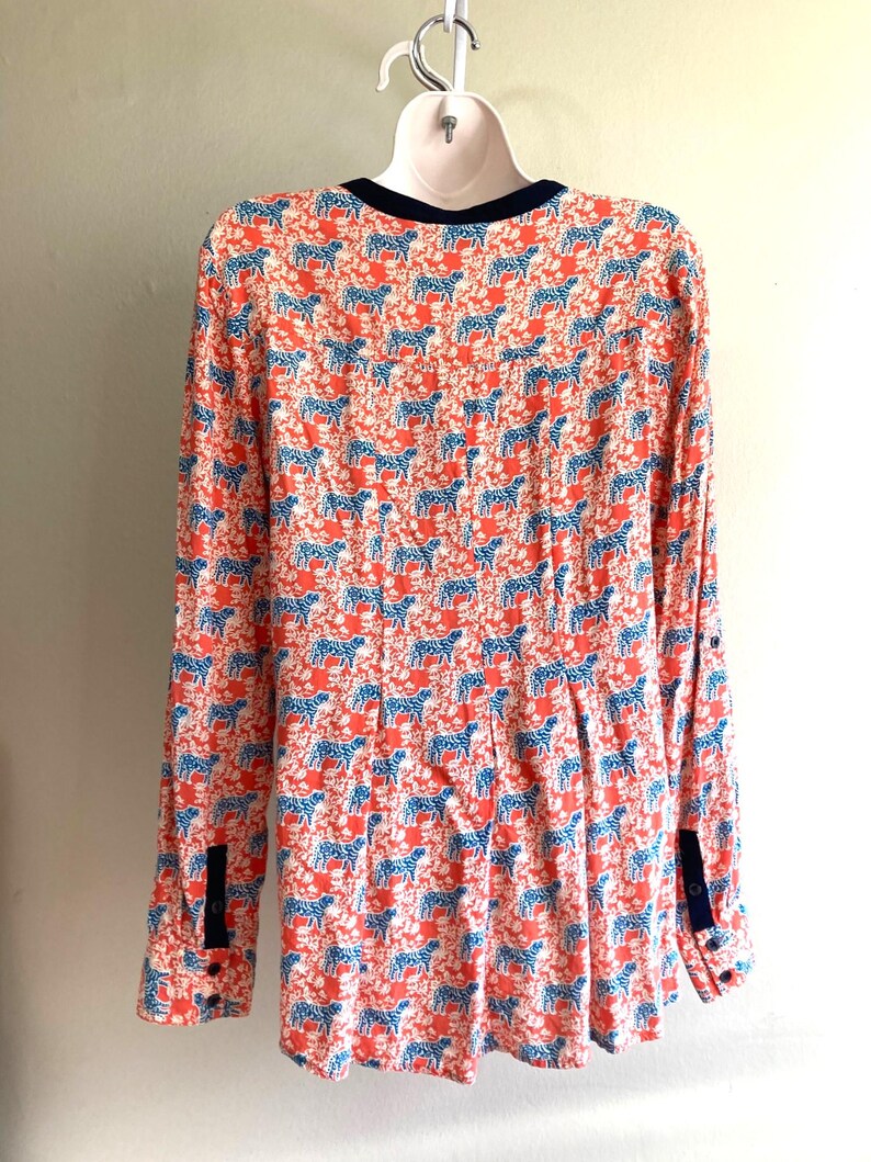 May include: A long-sleeved blouse with a vibrant pattern of blue zebras on a coral background. The blouse features a navy blue trim on the collar and cuffs, and is hanging on a white hanger.