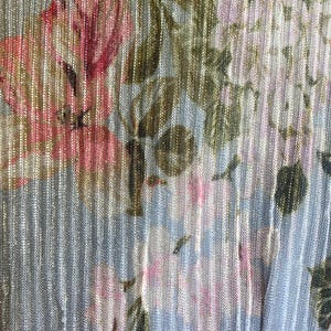 Op de afbeelding: Close-up van een stof met een bloemenpatroon. Het ontwerp toont roze en witte bloemen met groene bladeren op een lichtblauwe achtergrond. De stof heeft een verticaal gestreepte textuur met een metallic glans.