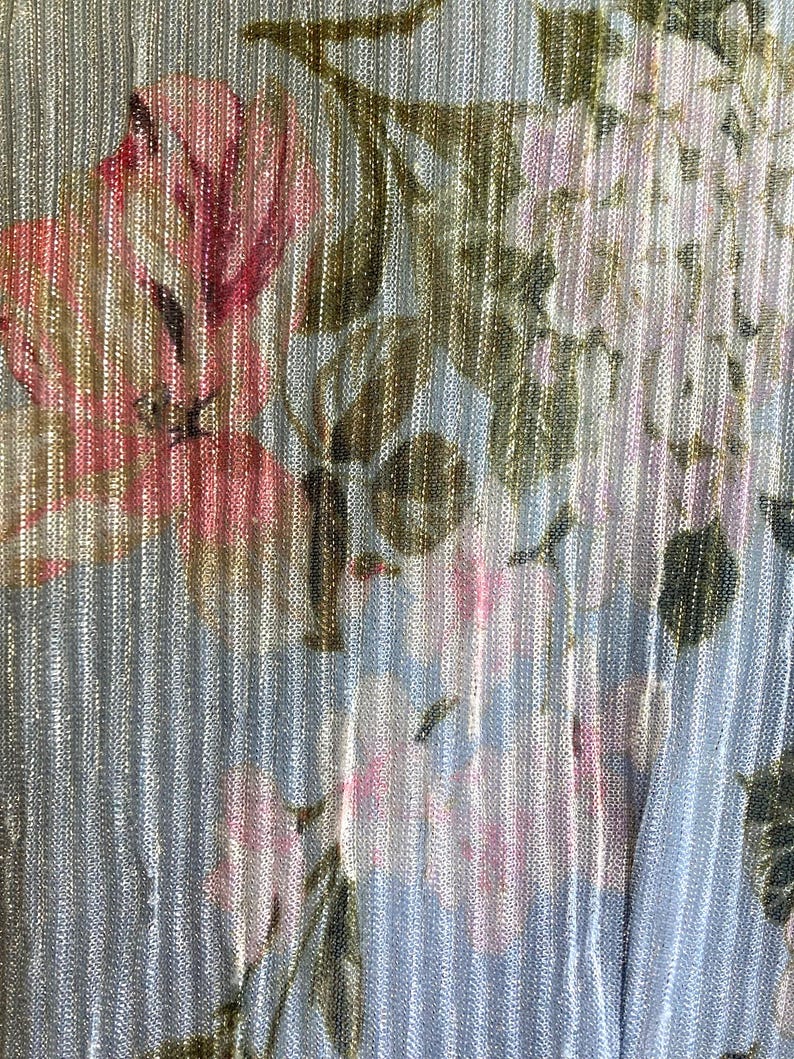 Op de afbeelding: Close-up van een stof met een bloemenpatroon. Het ontwerp toont roze en witte bloemen met groene bladeren op een lichtblauwe achtergrond. De stof heeft een verticaal gestreepte textuur met een metallic glans.
