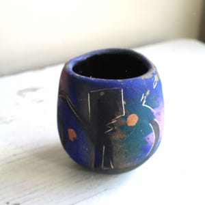 Puede incluir: Una pequeña maceta de cerámica azul con diseños abstractos en negro, blanco y naranja. La maceta tiene una forma redondeada y un acabado liso y brillante.