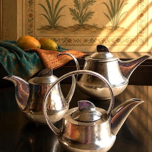 May include: Three silver-toned metal teapots with rounded bodies, curved spouts, and domed lids with finials. The teapots feature polished surfaces with visible metallic reflections and are arranged on a dark surface, showcasing their distinct shapes.