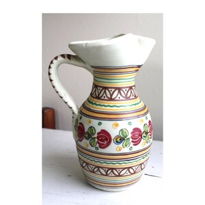 May include: A white ceramic pitcher with a wide mouth and a handle. The pitcher is decorated with hand-painted red roses, green leaves, and yellow, brown, and green stripes.
