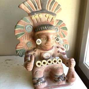 Op de afbeelding: Een terracotta-kleurig keramieken beeldje van een zittende figuur met een uitgebreide hoofdtooi met groene en beige accenten. Het figuur heeft ronde oorornamenten, een centrale borstschijf en vijf kleine ringen over de taille.