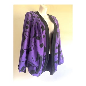 Può includere: Un cardigan viola aperto sul davanti con un motivo geometrico nero di cerchi, triangoli e rettangoli. Il capo presenta maniche lunghe, un bordo nero a tinta unita lungo l'apertura frontale e due tasche anteriori integrate nel tessuto.