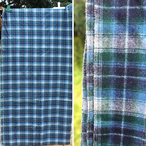 Puede incluir: Un textil rectangular con un patrón de cuadros tejido en tonos azul, verde, blanco y azul marino oscuro. La tela muestra un diseño de cuadrícula consistente con líneas horizontales y verticales que se cruzan, exhibiendo una superficie texturizada.