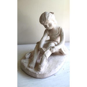 May include: A cream-coloured sculpture of a young girl seated, tying her ballet shoe. The figure is wearing a leotard and a skirt. The sculpture is on a rounded base and has a matte finish.