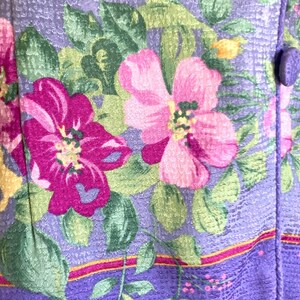 May include: Close-up of a floral fabric with a lavender background. The fabric features large pink and magenta flowers with green leaves and yellow accents. A purple button is visible on the right side, suggesting a garment or textile.