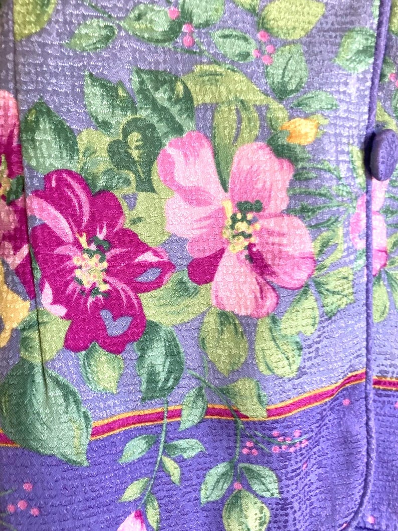May include: Close-up of a floral fabric with a lavender background. The fabric features large pink and magenta flowers with green leaves and yellow accents. A purple button is visible on the right side, suggesting a garment or textile.