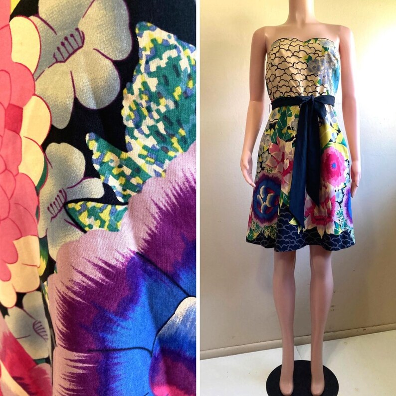 May include: A strapless knee-length dress featuring a vibrant floral print with large pink, purple, and blue blossoms on a black background. The bodice has a scalloped pattern, and a solid black sash ties at the waist, cinching the skirt.